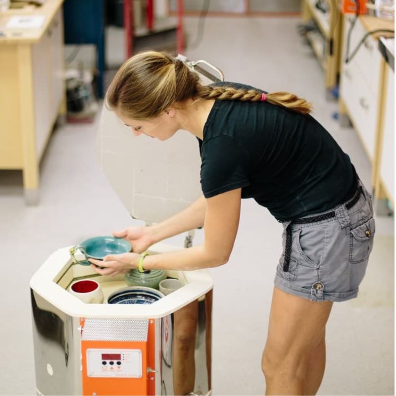 Woman in gray shorts - black shirt loading glazed ceramic pieces in to an open EMERGE ceramic-pottery kiln (model AF3C 15/13) from Jen-Ken  with the standard Orton AutoFire AF3C 3-Button Controller.